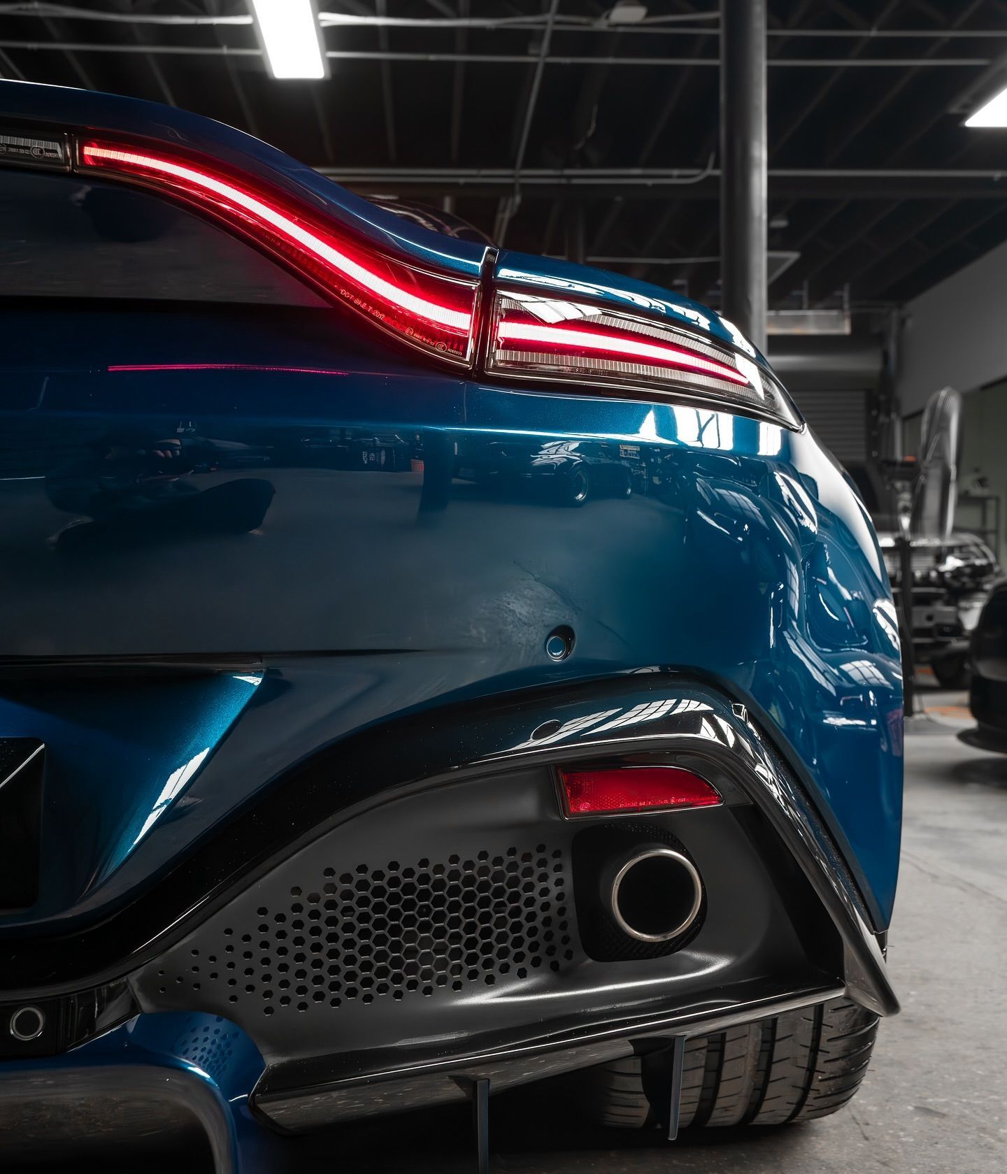 Blue Aston Martin car rear detail, showing taillight, exhaust, and honeycomb grille in a garage.