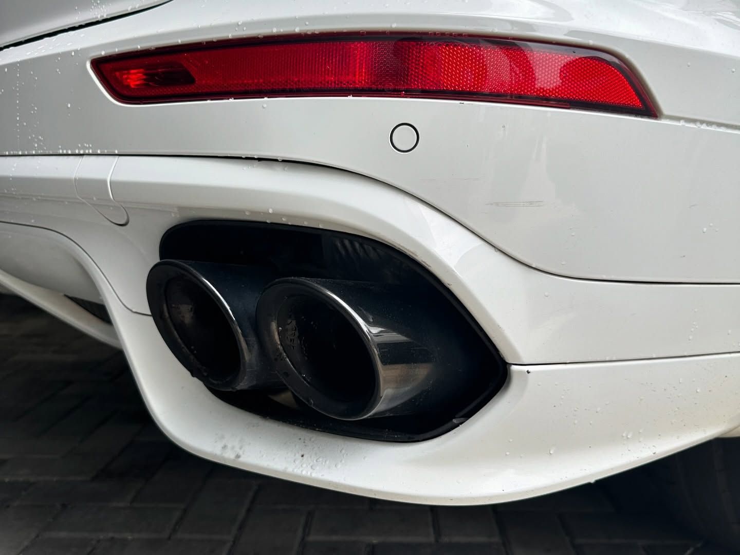 Close-up of a white car's rear bumper, featuring two black exhaust pipes, a red taillight, and a parking sensor.