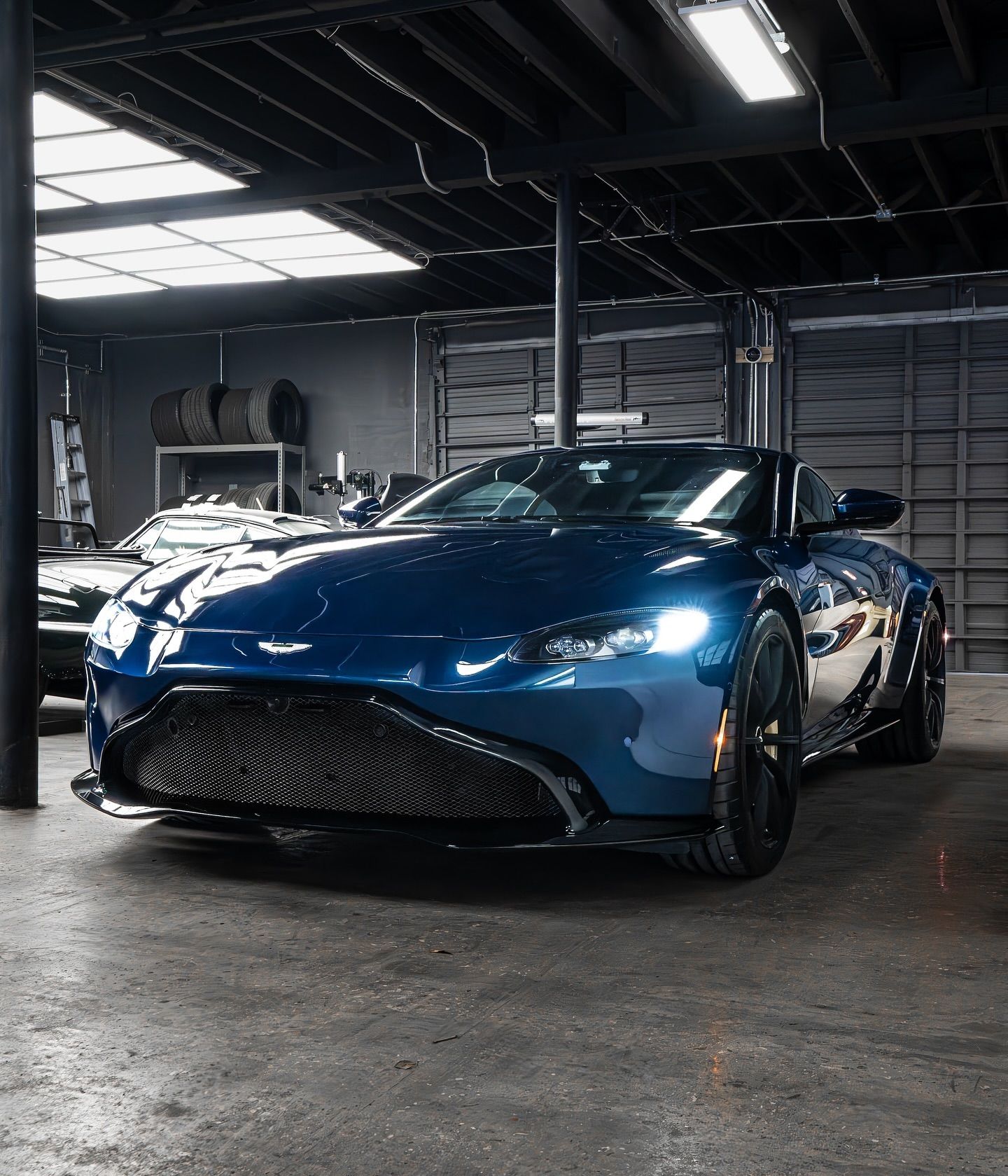 Dark blue Aston Martin sports car in a garage with black trim, under overhead lighting.