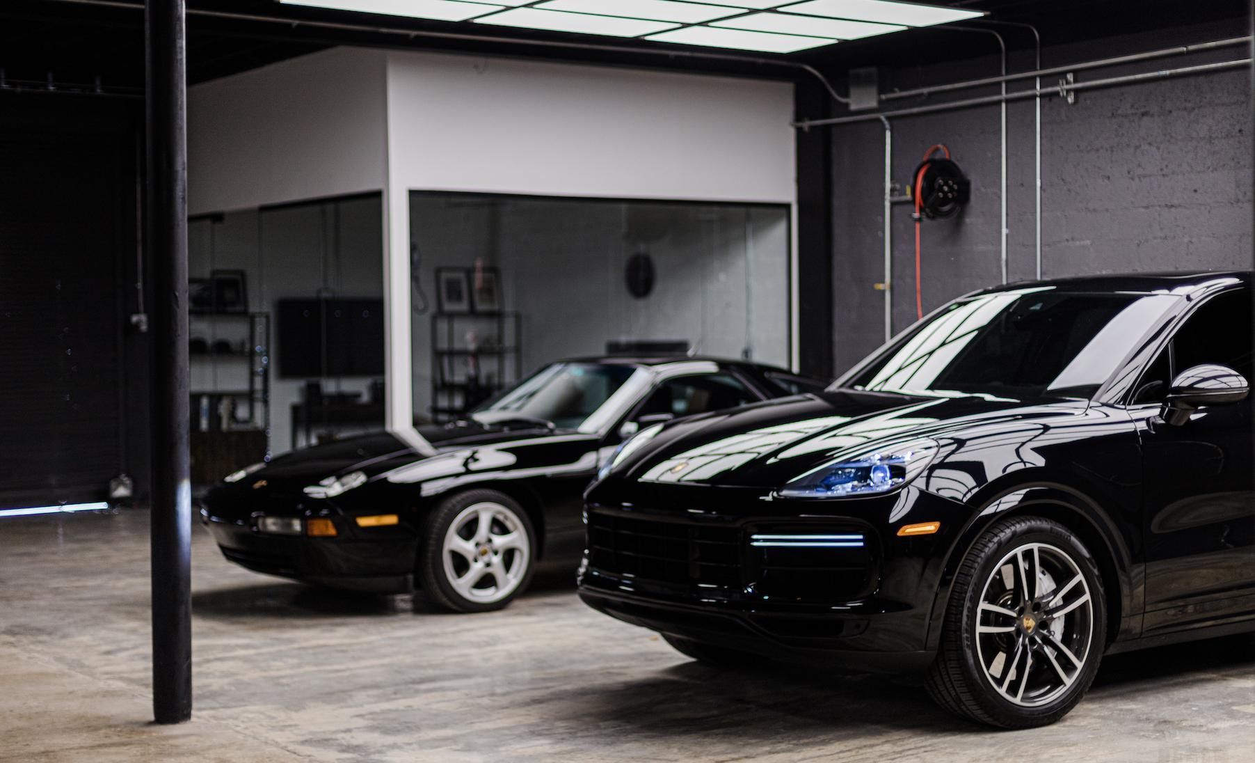 Two black Porsches in a garage. One is a classic model, the other is modern. A glass-walled office is in the background.