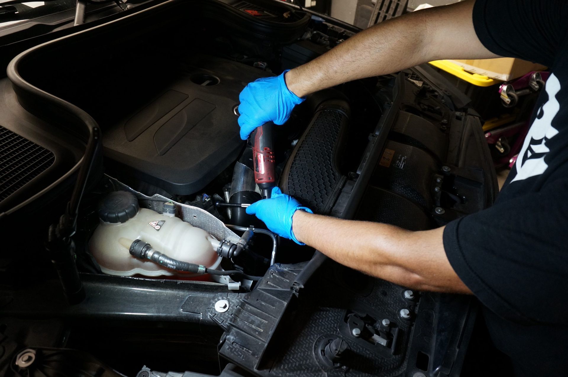 Person in blue gloves working on a car engine bay, using a power tool near a coolant reservoir.