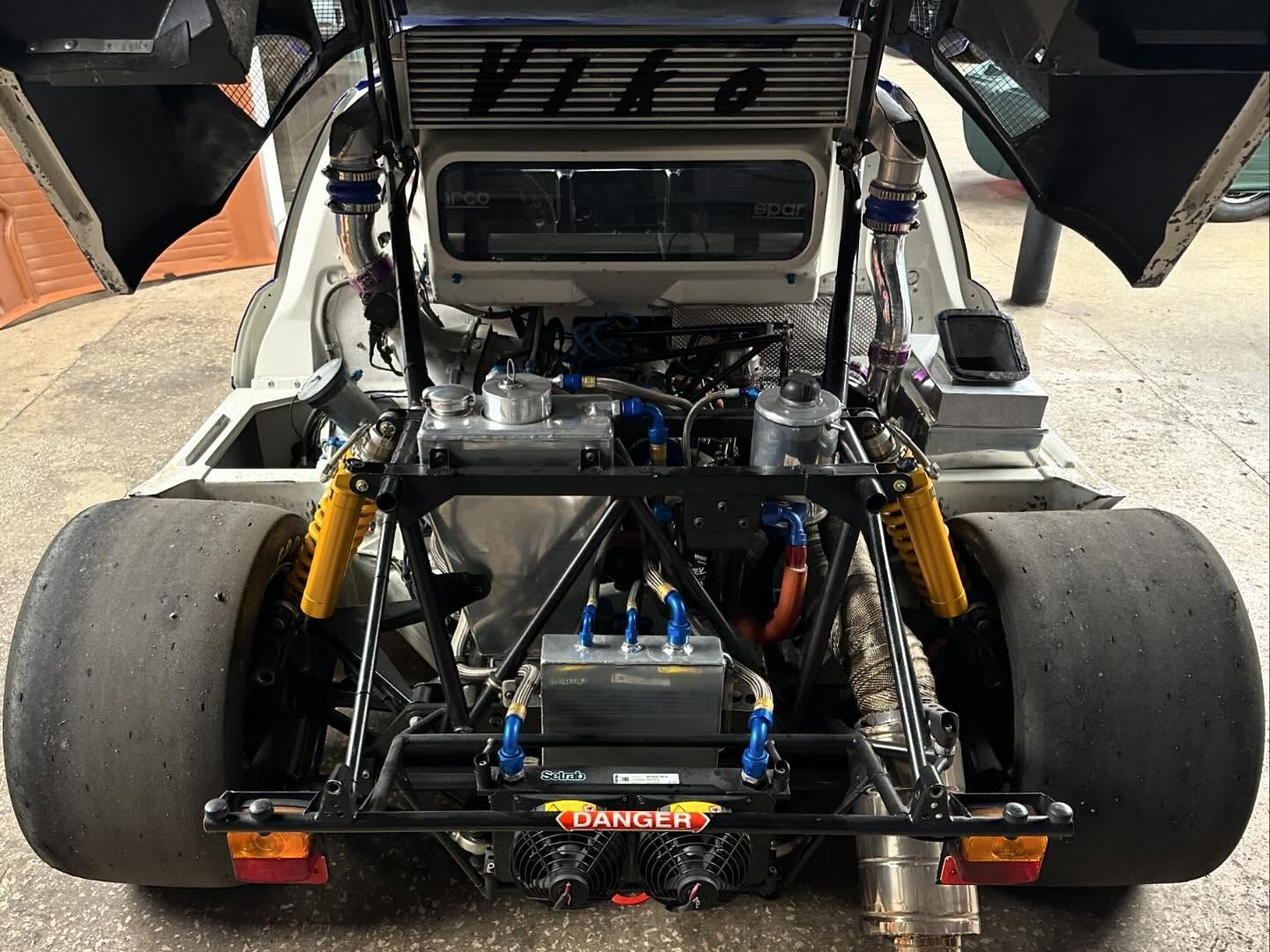 Rear view of a white race car with exposed engine, open doors, and large tires.
