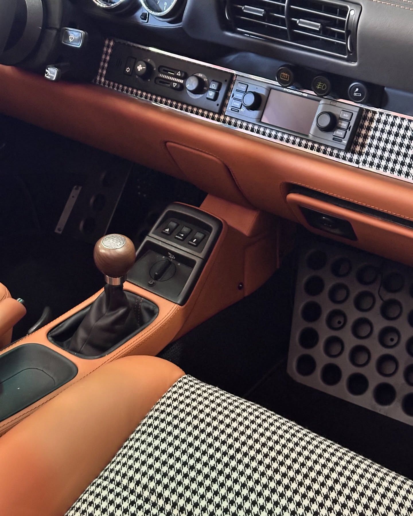 Interior of a tan and black car, gear shift, patterned fabric, and circular foot pedals.