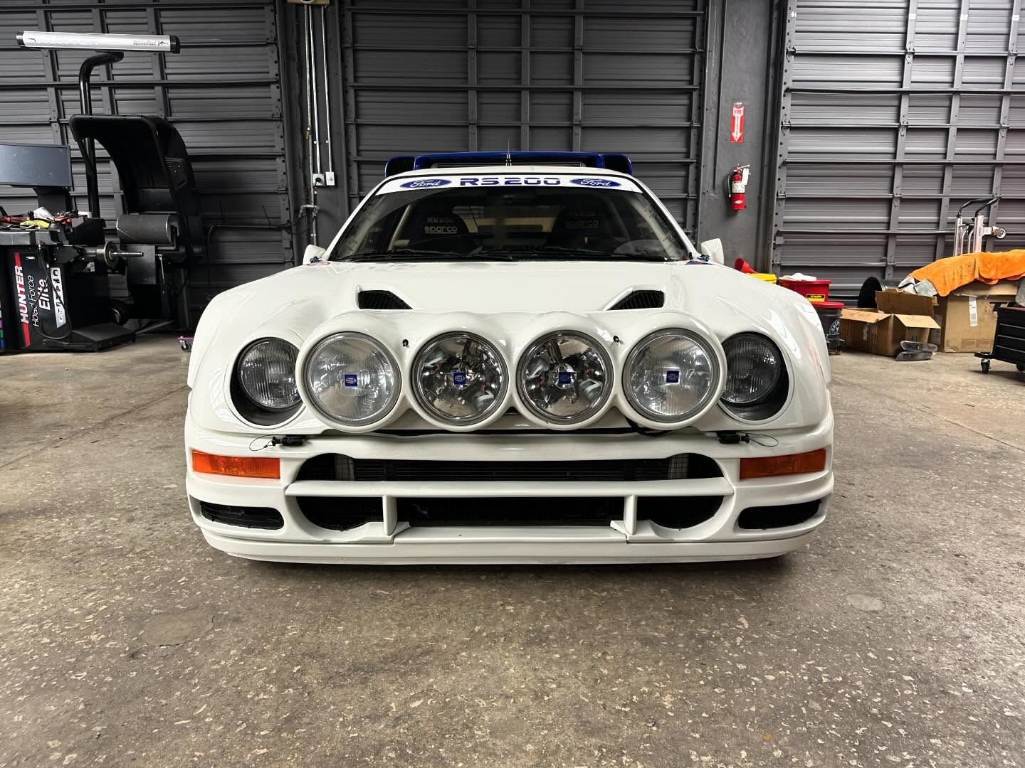 White rally car with extra headlights, parked inside a garage.