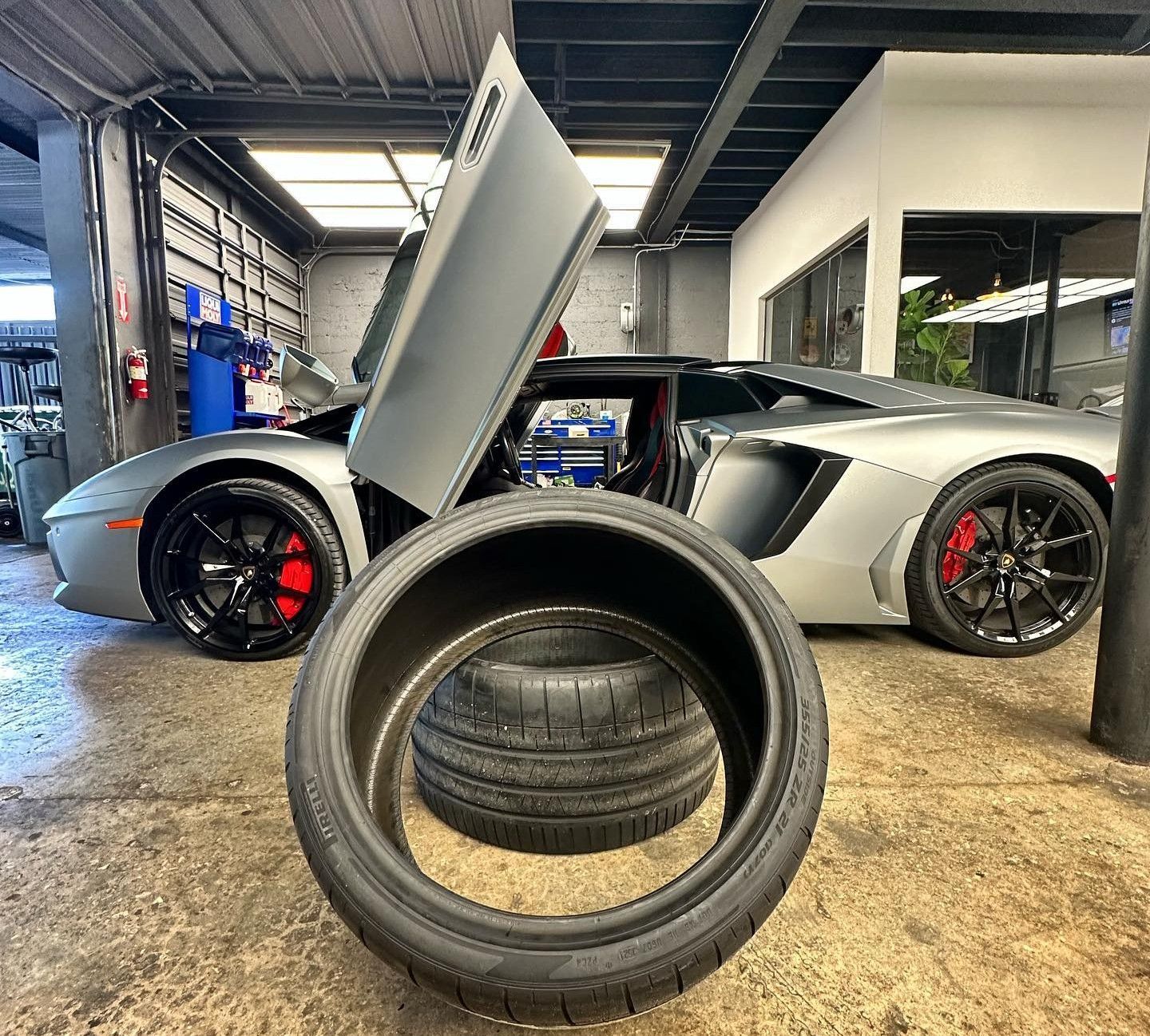 Silver Lamborghini with open door in a garage, tires in foreground.