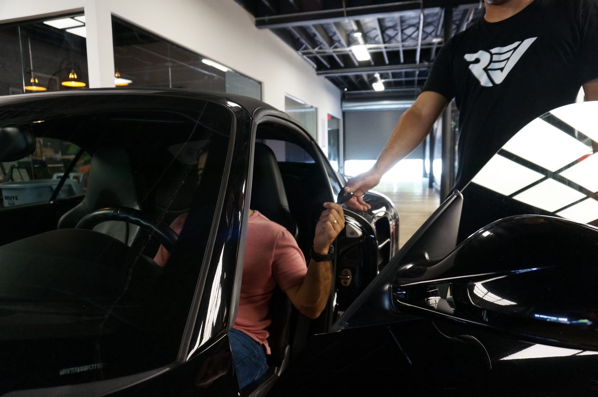 Man getting into a black car; another man holds the door open. Inside a showroom.