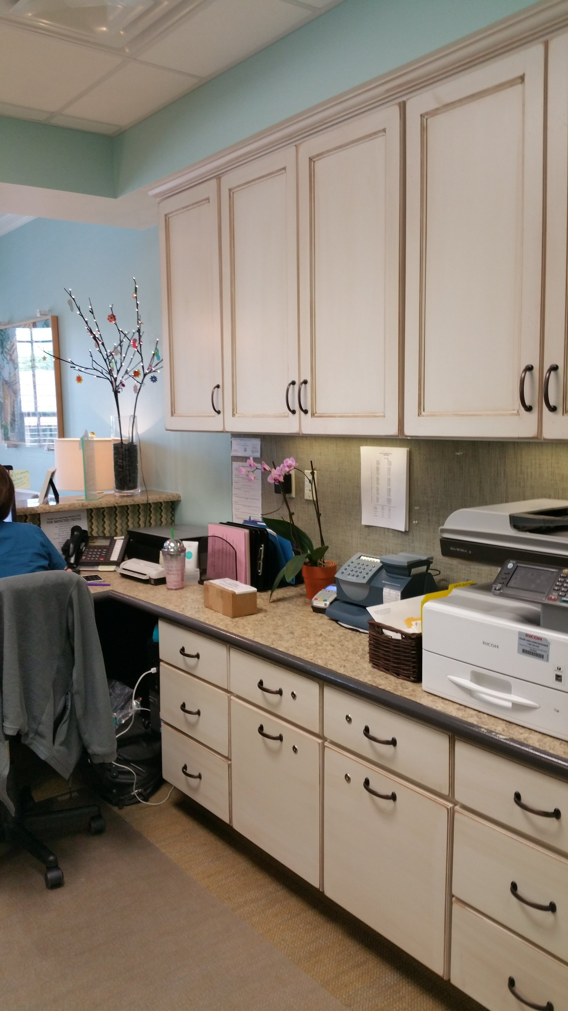 A woman is sitting at a desk in a kitchen with white cabinets.
