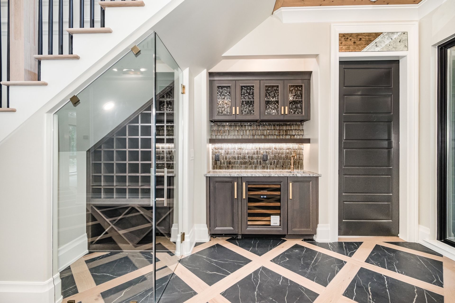 Wine cellar under stairs with a glass door, and a bar with cabinets, tiled floor.