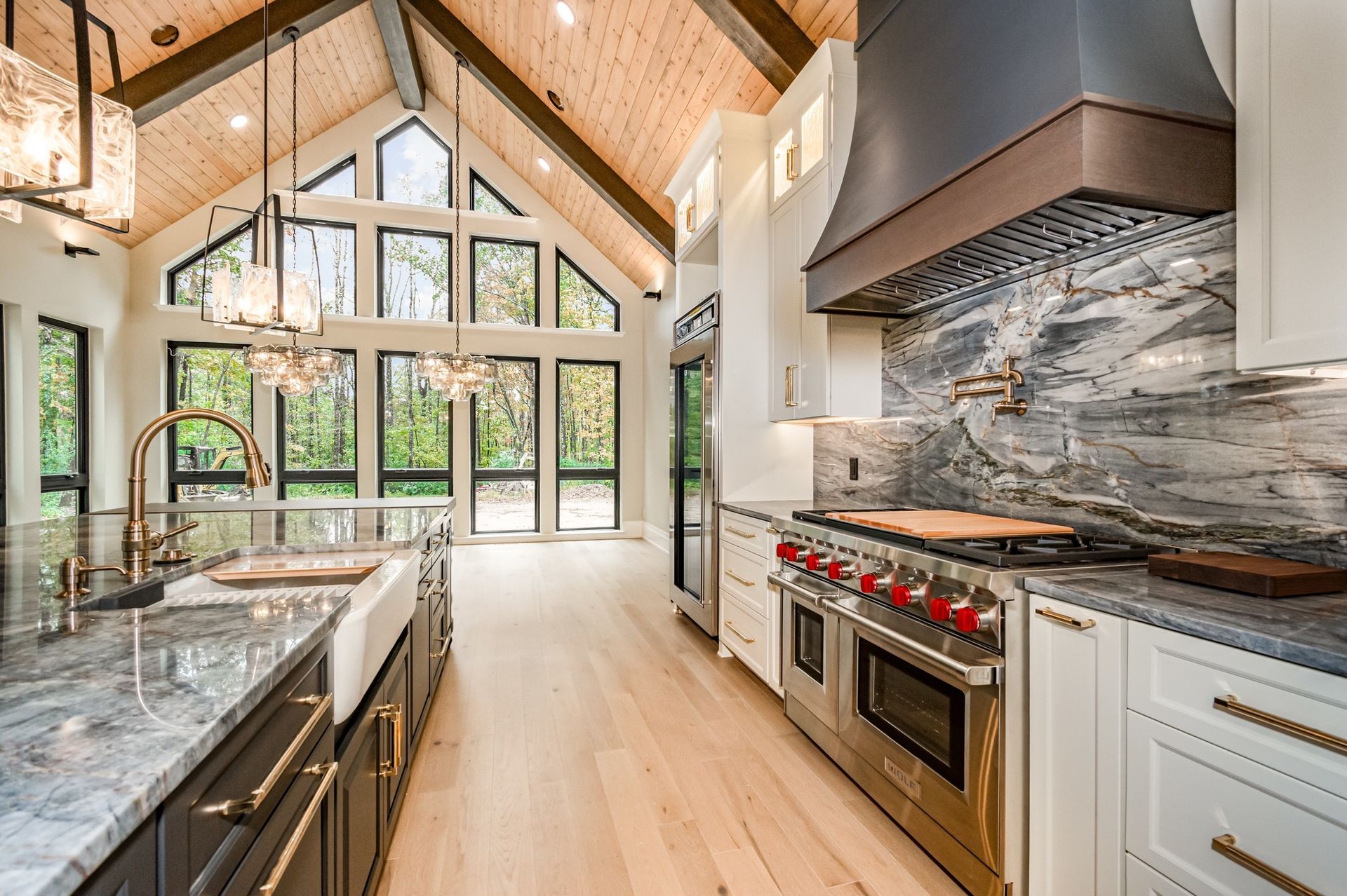 A modern kitchen with wood ceiling and floors, a large island, stainless steel appliances, and large windows looking out onto trees.
