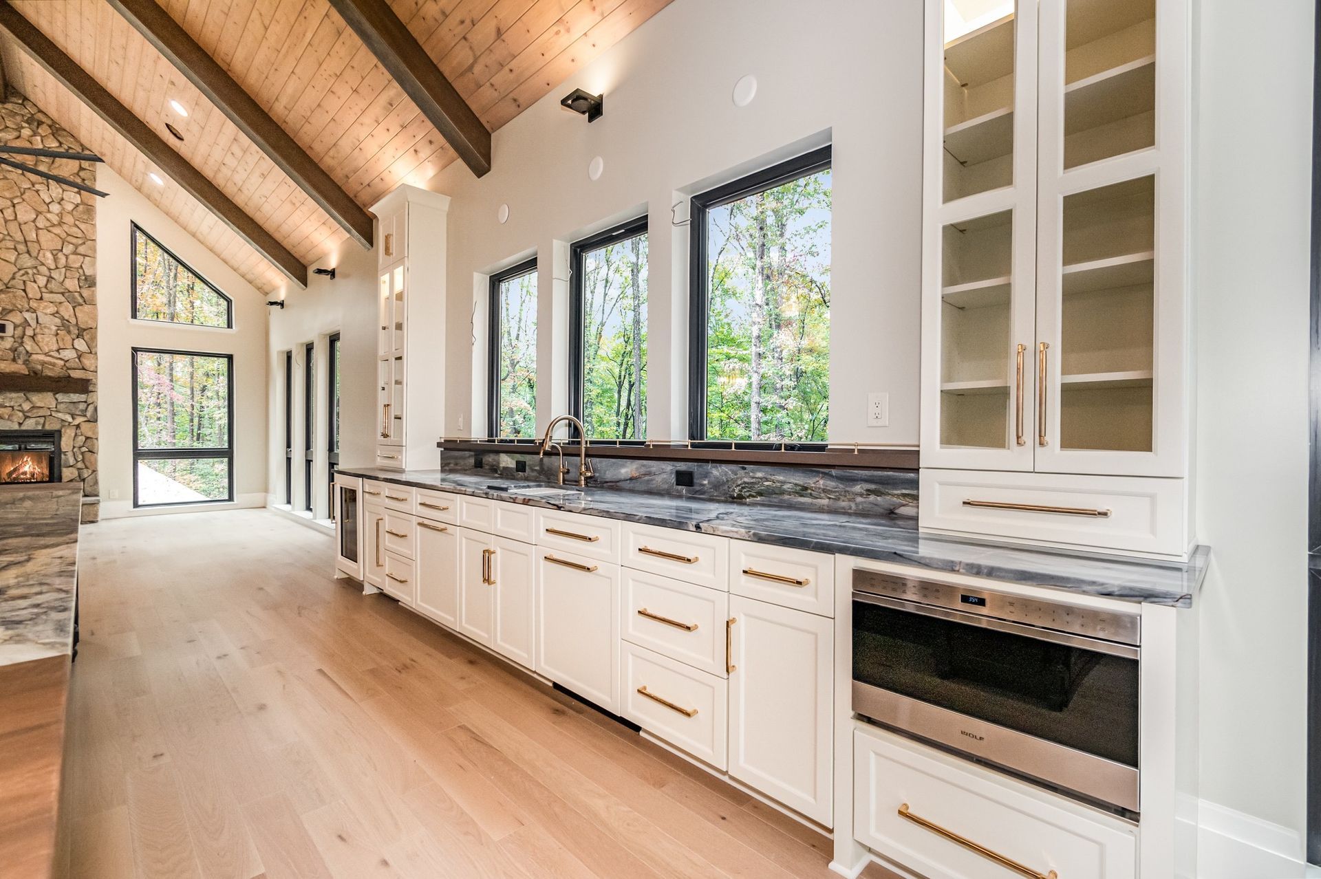 Modern kitchen with white cabinets, wood ceiling, and large windows overlooking trees.