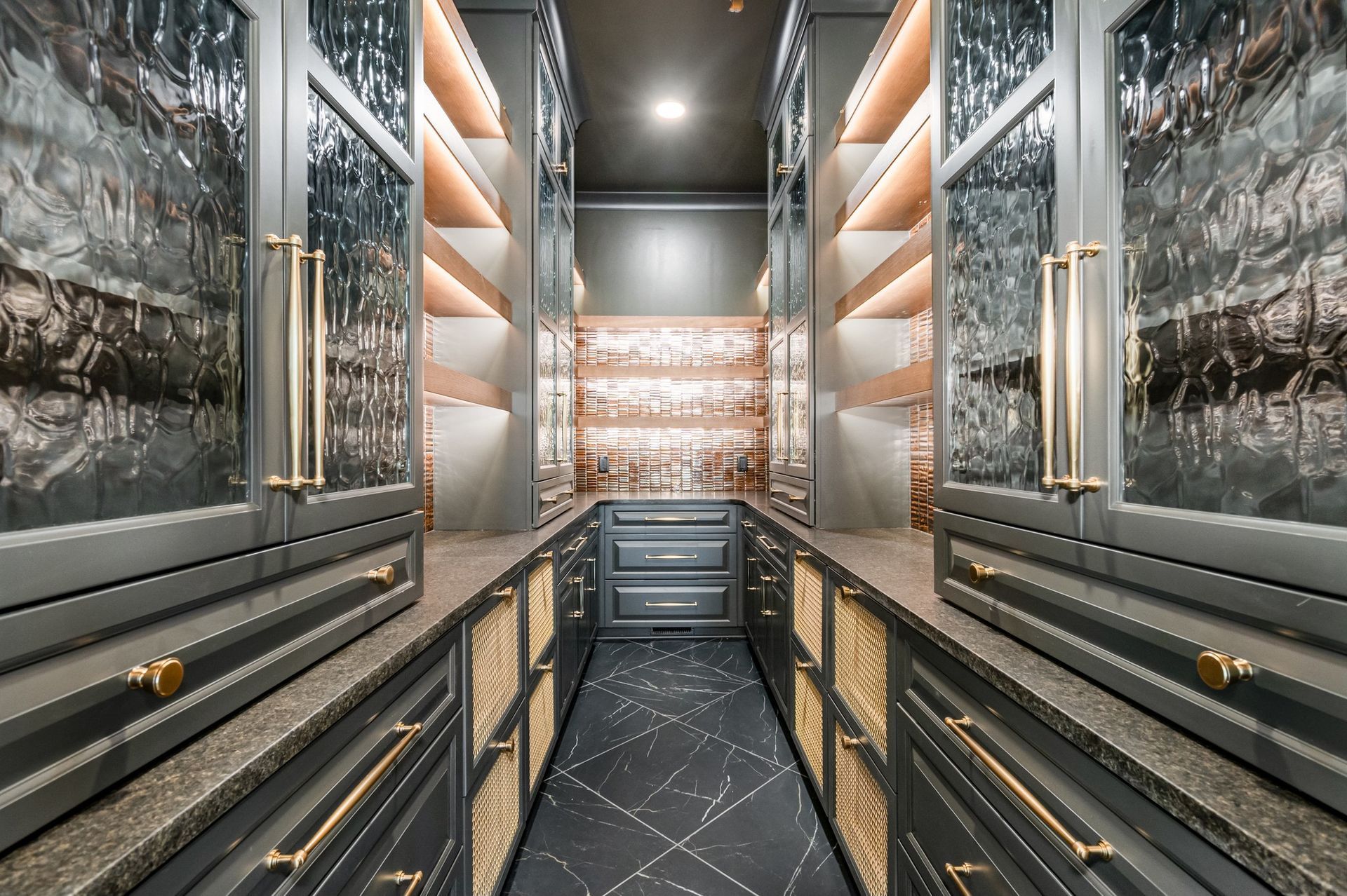 Walk-in bar with gray cabinets, gold accents, and glass shelves filled with liquor bottles and glassware.