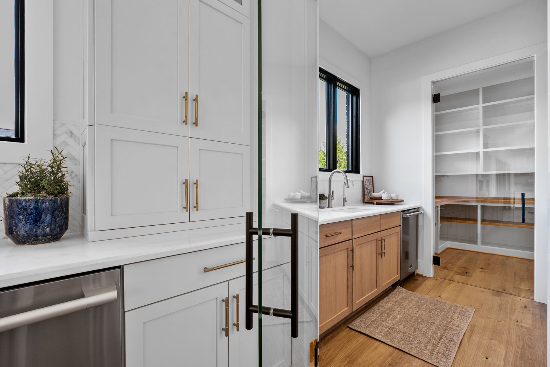 Laundry room with white cabinets, a sink, and pantry. Black window frame, wooden floor, and rug.