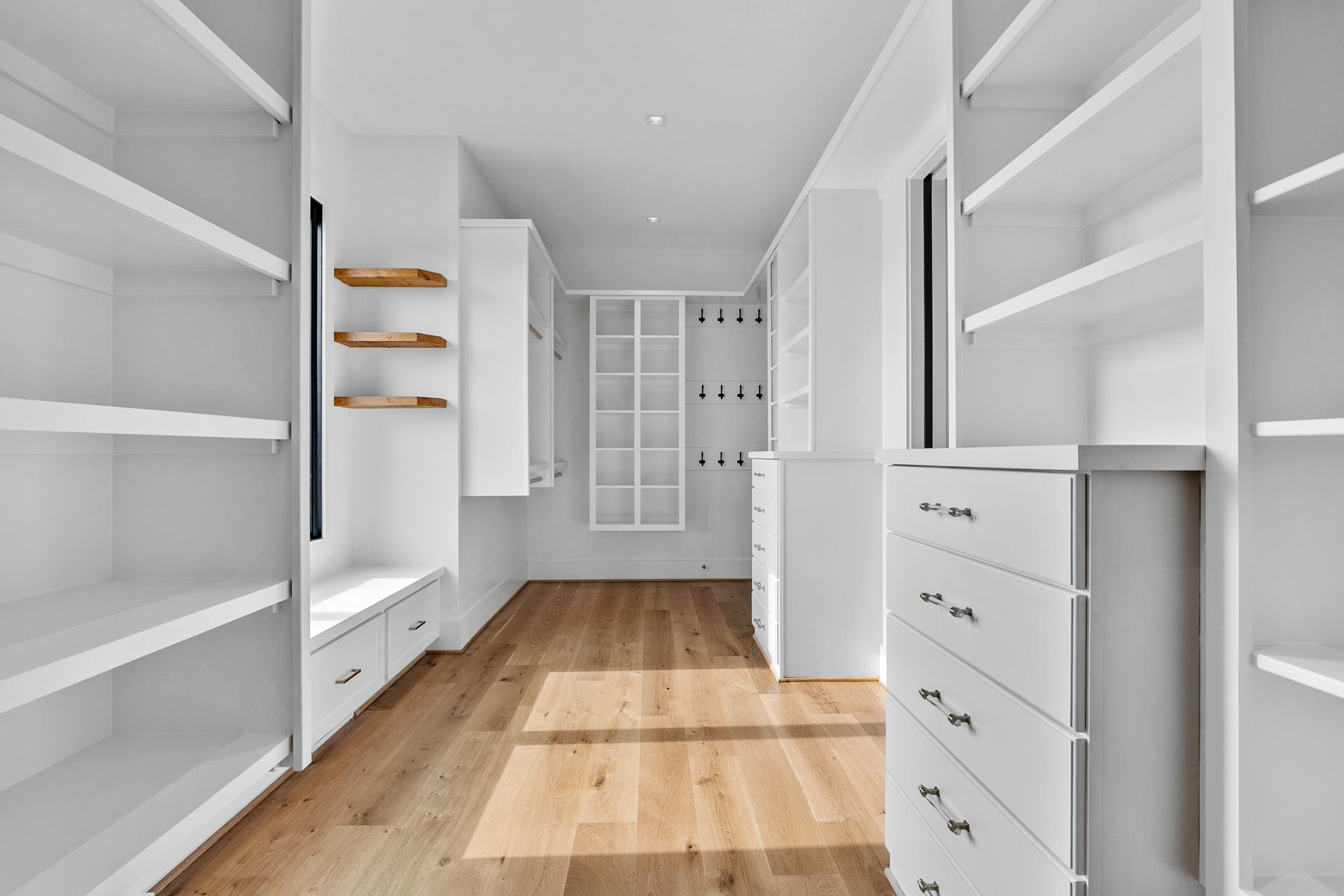 Empty, white-shelved walk-in closet with wooden floor. Cabinets, drawers, shelves, and a bench are visible.