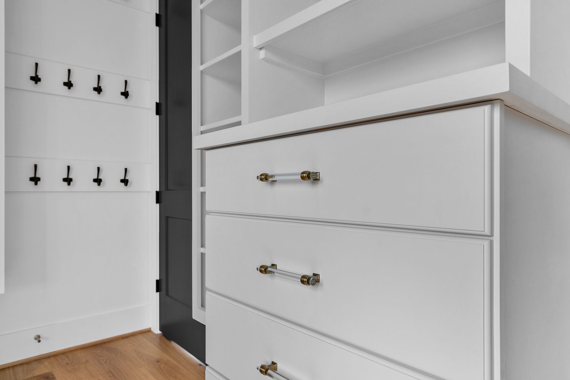 White custom closet drawers with clear and brass pulls, adjacent to shelves and hook rack.