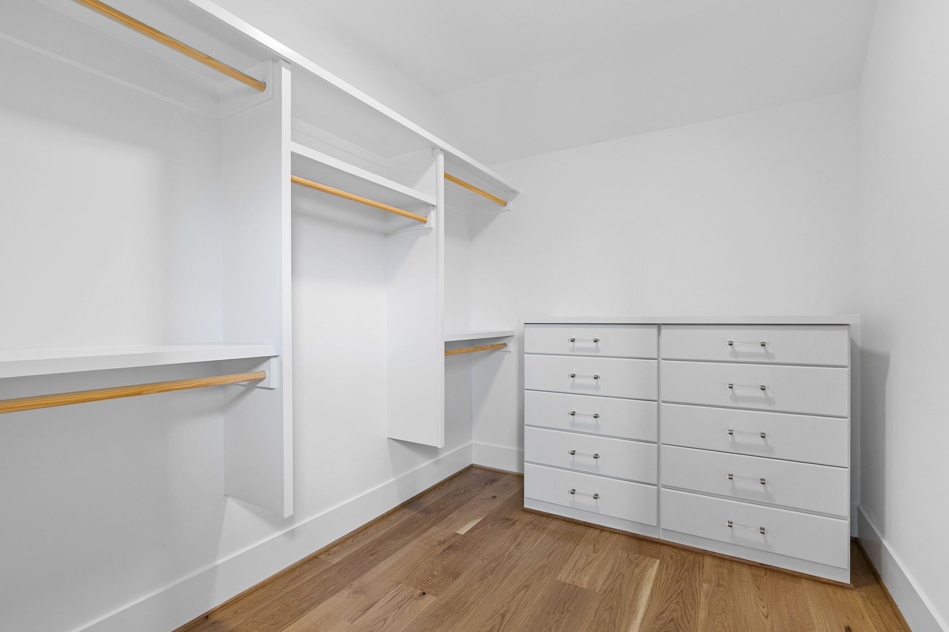 Empty white walk-in closet with wooden shelves and a dresser on a hardwood floor.