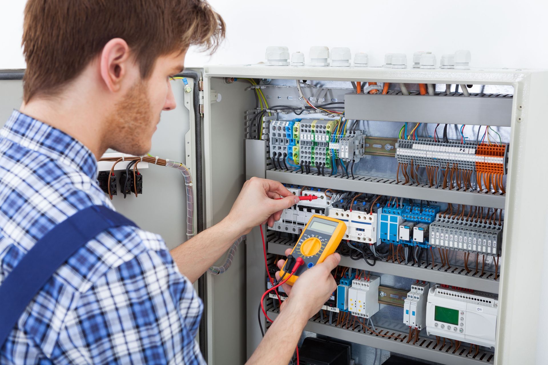 Electrician in a blue plaid shirt, inspecting wiring in a gray electrical panel with a yellow multimeter.