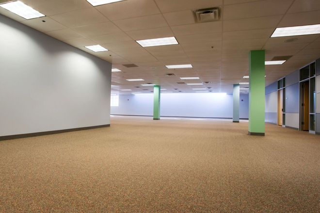 Empty office space with beige carpet, white walls, and green support columns under fluorescent ceiling lights.