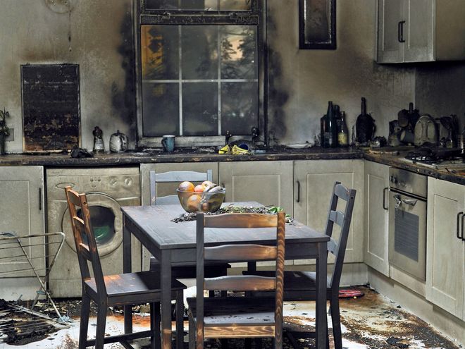 Burned kitchen with soot-stained walls, charred cabinets, and a dining table amid fire damage.