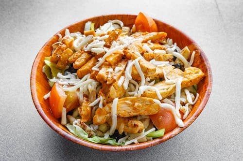 A bowl of food with chicken , cheese and tomatoes on a table.