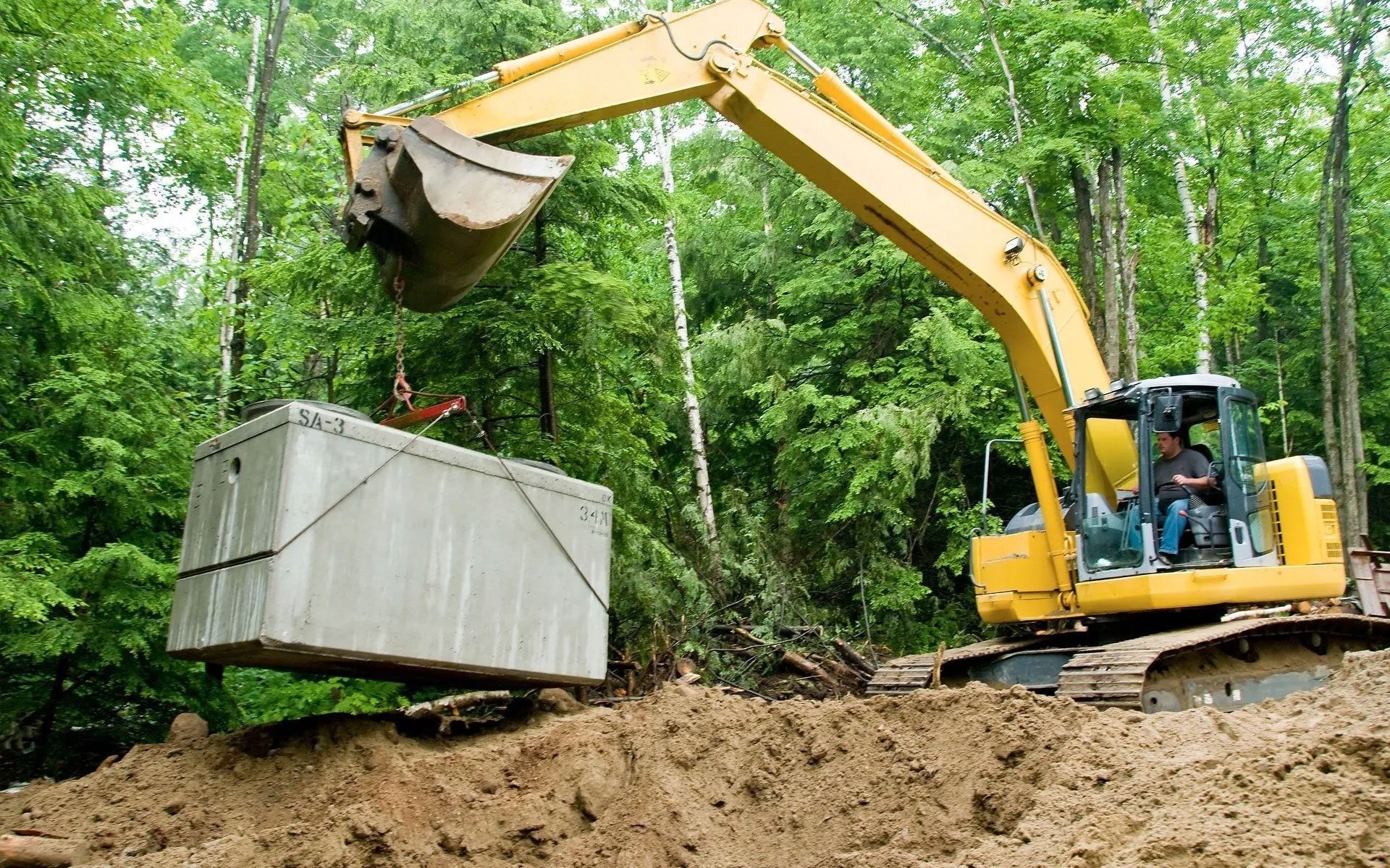 Yellow excavator lifting a large concrete block in a wooded construction site