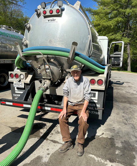 Man sitting in front of a large vacuum truck with green hoses at a roadside work site