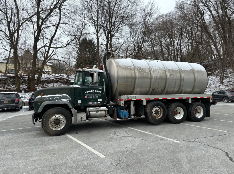 Green tanker truck parked on a snowy lot with bare trees in the background