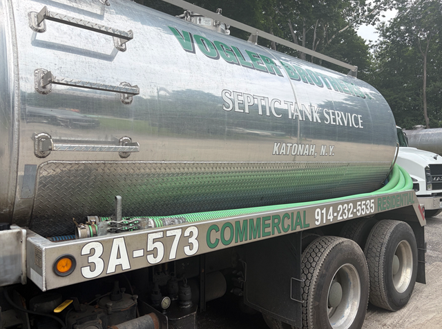 Green commercial septic service tanker truck with white lettering parked outdoors under trees