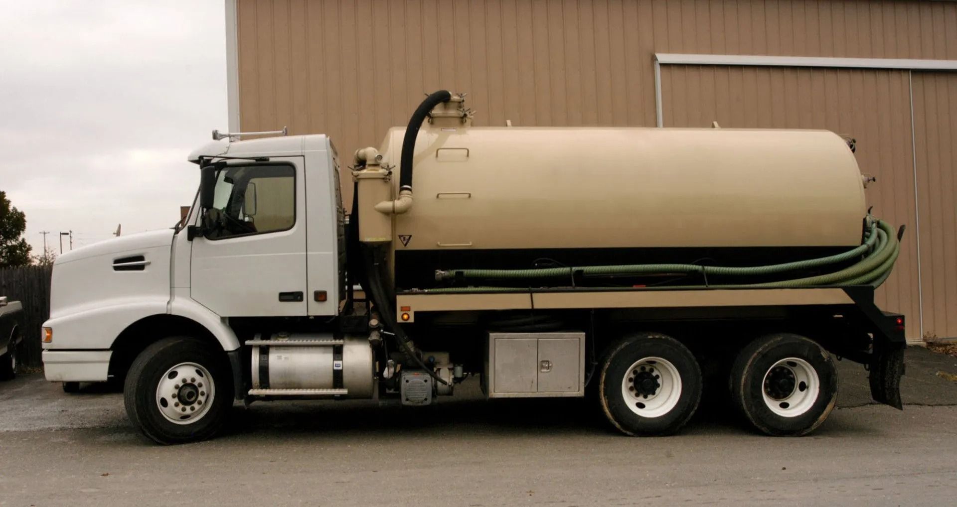 White tanker truck parked beside a tan warehouse on a cloudy day