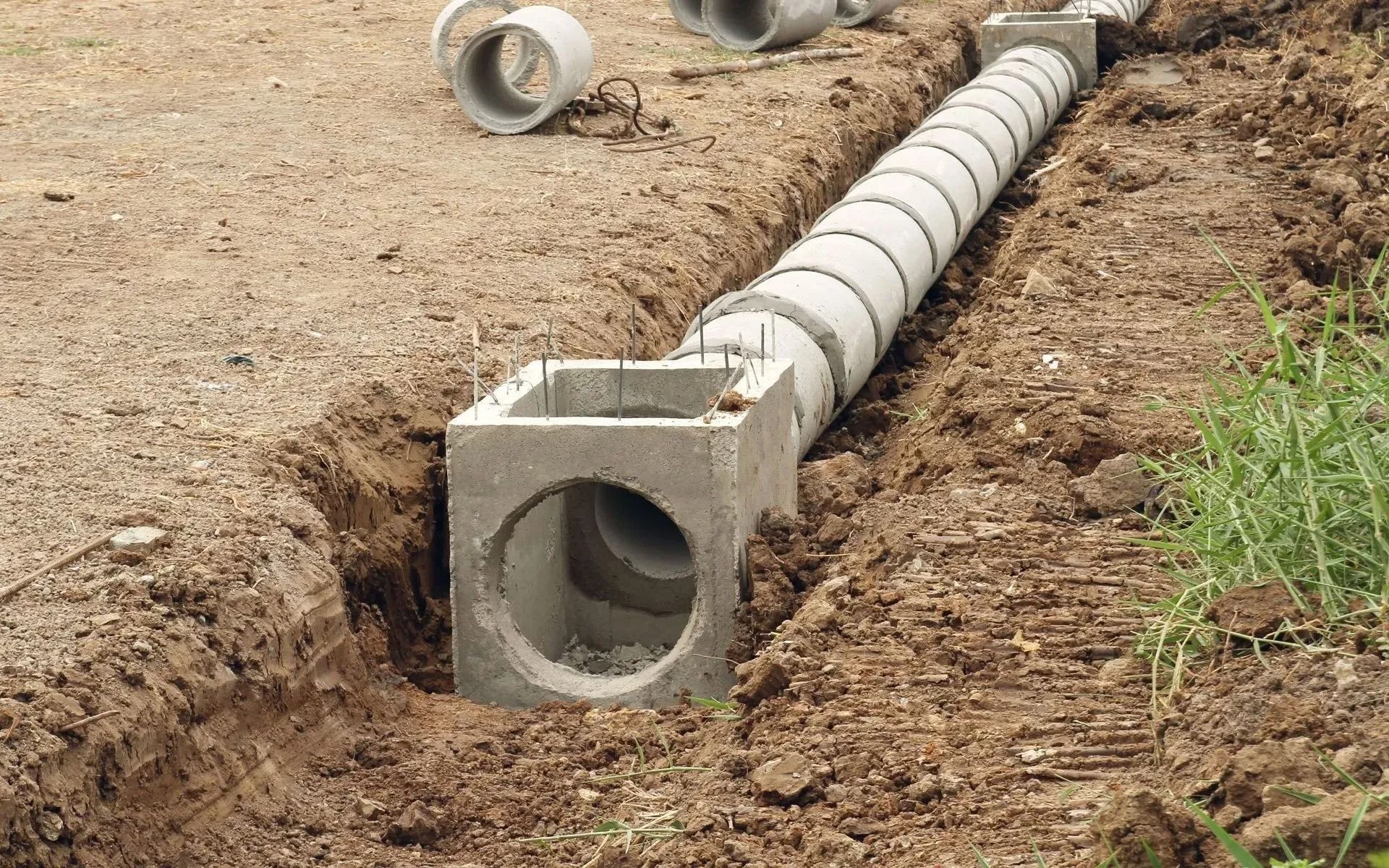 Concrete utility conduit laid in a trench on bare soil, with a square access box in the foreground.