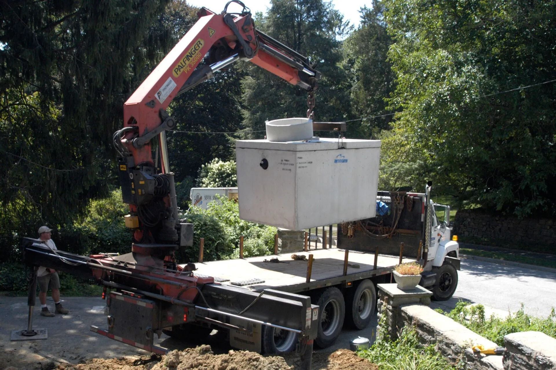 Crane lifting a white utility box onto a flatbed truck in a wooded roadside area