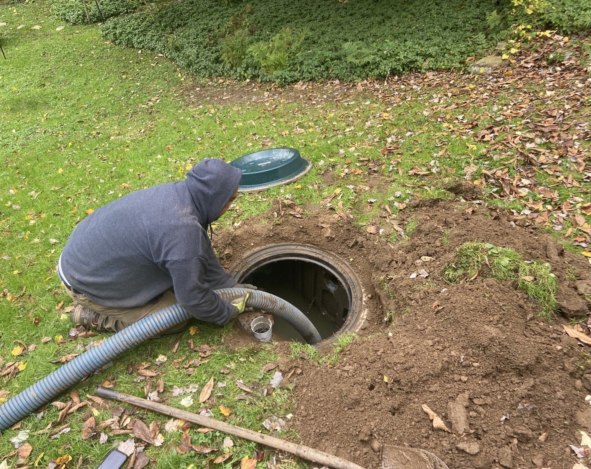 Person in a gray hoodie vacuuming a round underground opening in a grassy yard.