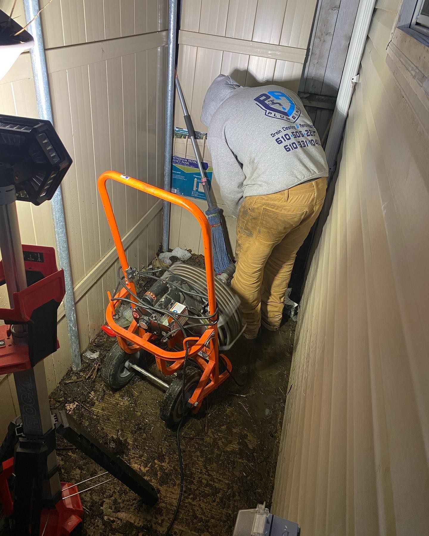Person in hoodie operating an orange pressure washer in a tight outdoor space between buildings.
