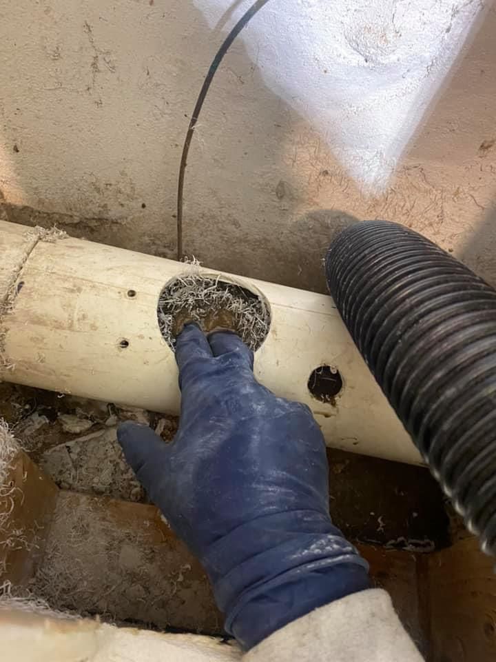 Gloved hand touching a hole in a white pipe, debris visible. Black hose nearby, in a cluttered, dusty area.