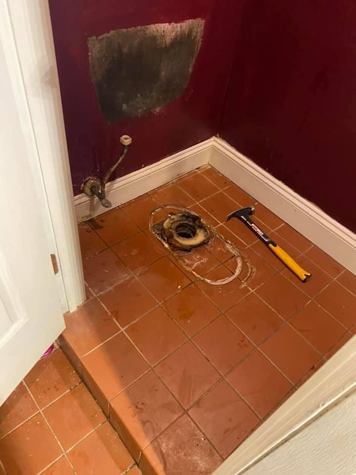 Bathroom under renovation; red tile floor, exposed toilet flange, damaged wall, hammer.