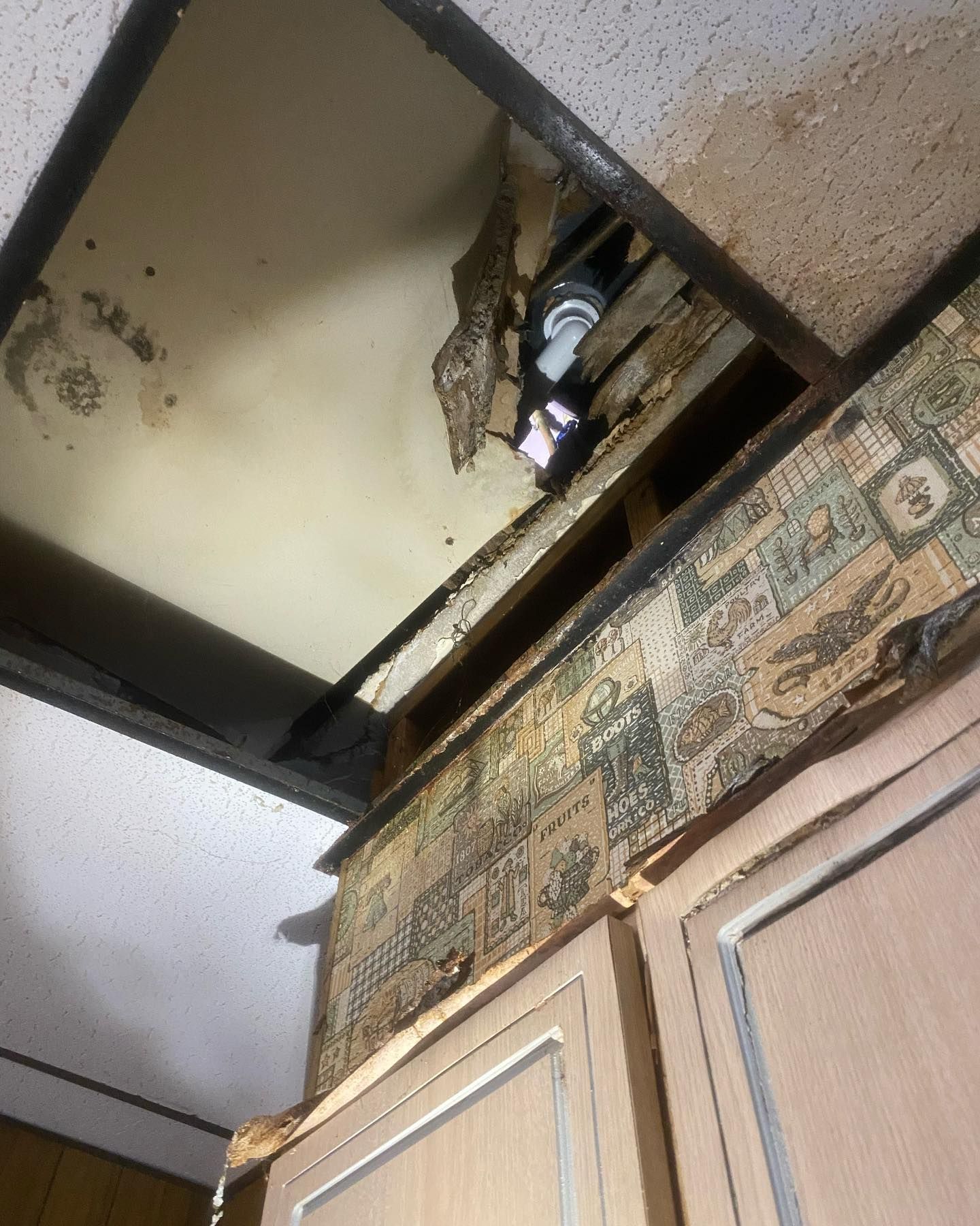 Damaged ceiling revealing wires and a light fixture above kitchen cabinets. Wallpaper peeling.