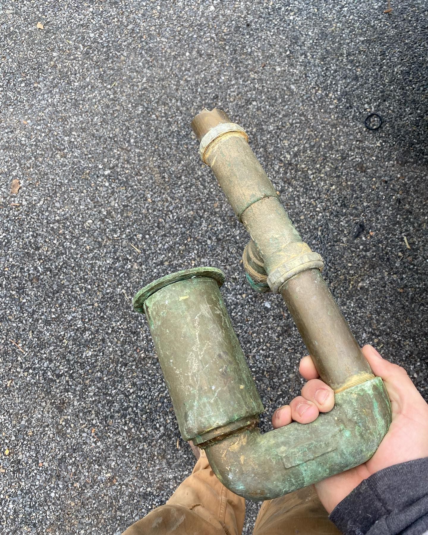 Person holding a corroded, copper drain pipe, likely from plumbing, outdoors on asphalt.