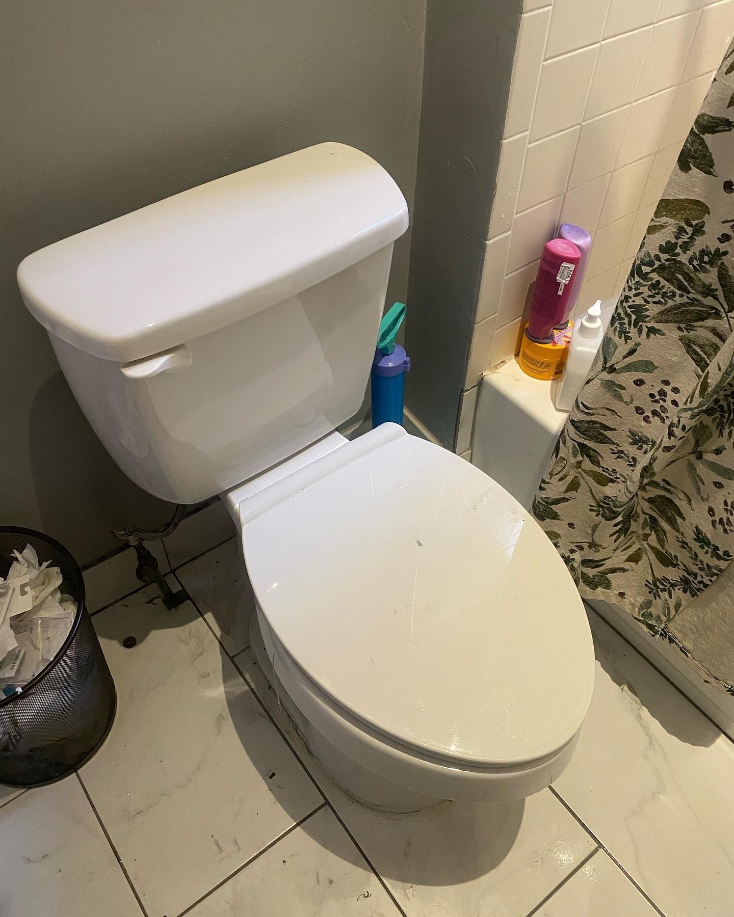 White toilet in a bathroom with a wastebasket, bottles, and a shower curtain.