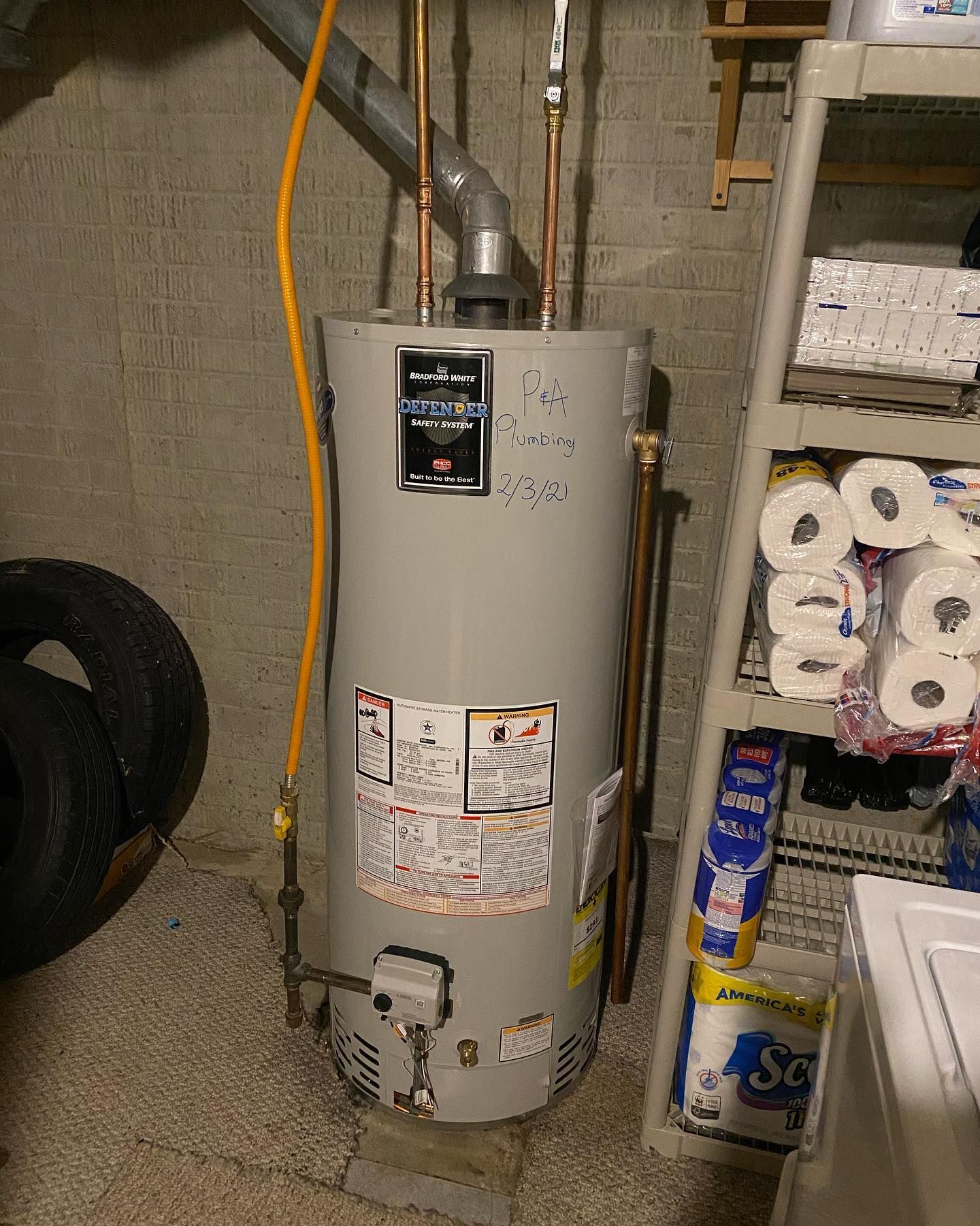 A water heater in a basement; gray tank, copper pipes, yellow gas line, with tires and shelves nearby.