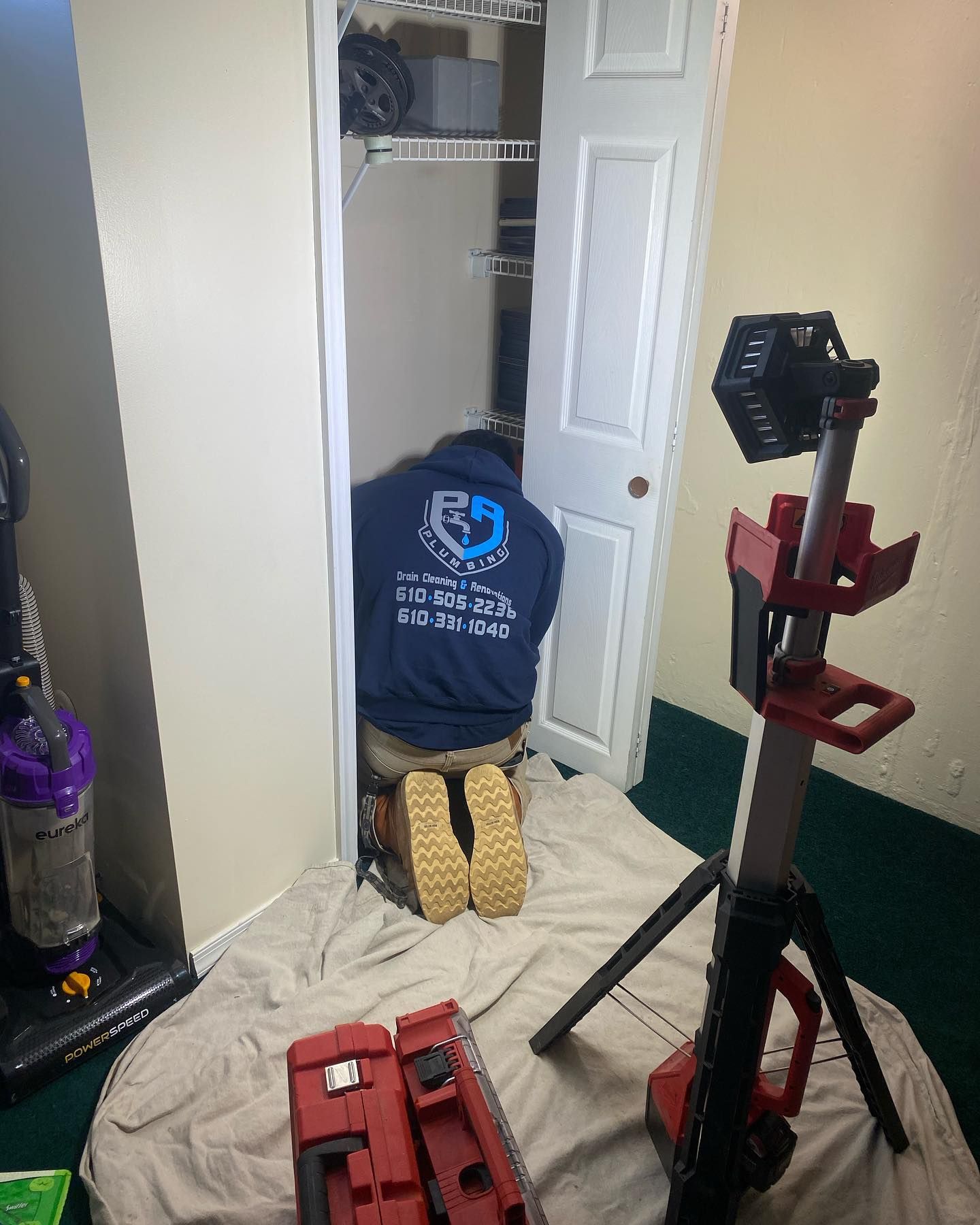 Person kneeling in closet, wearing work boots and a blue shirt. A work light and tool case are in the foreground.