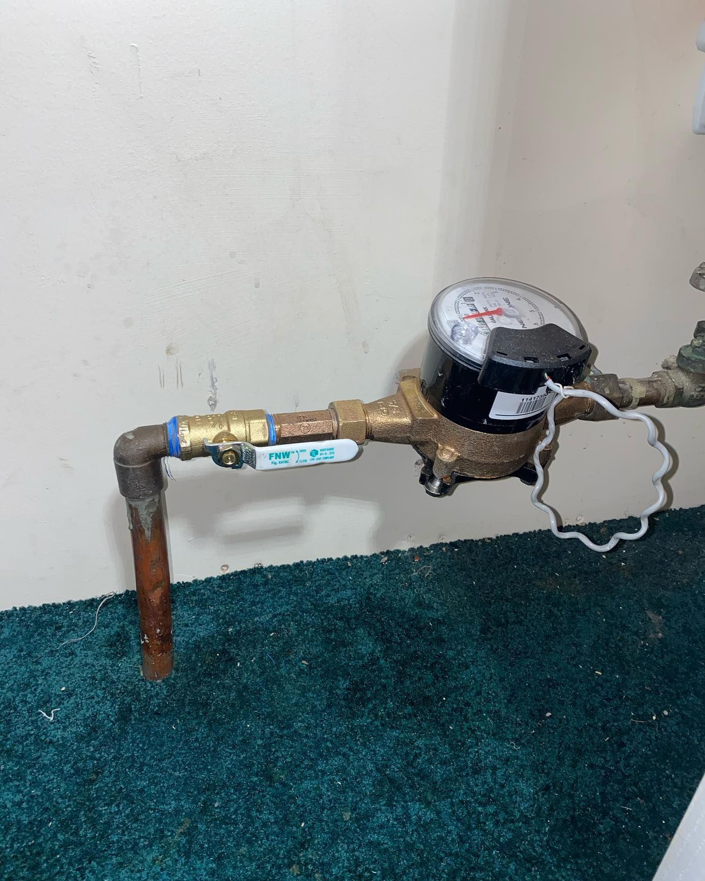 A water meter in a room with green carpet, pipes, and a gauge.