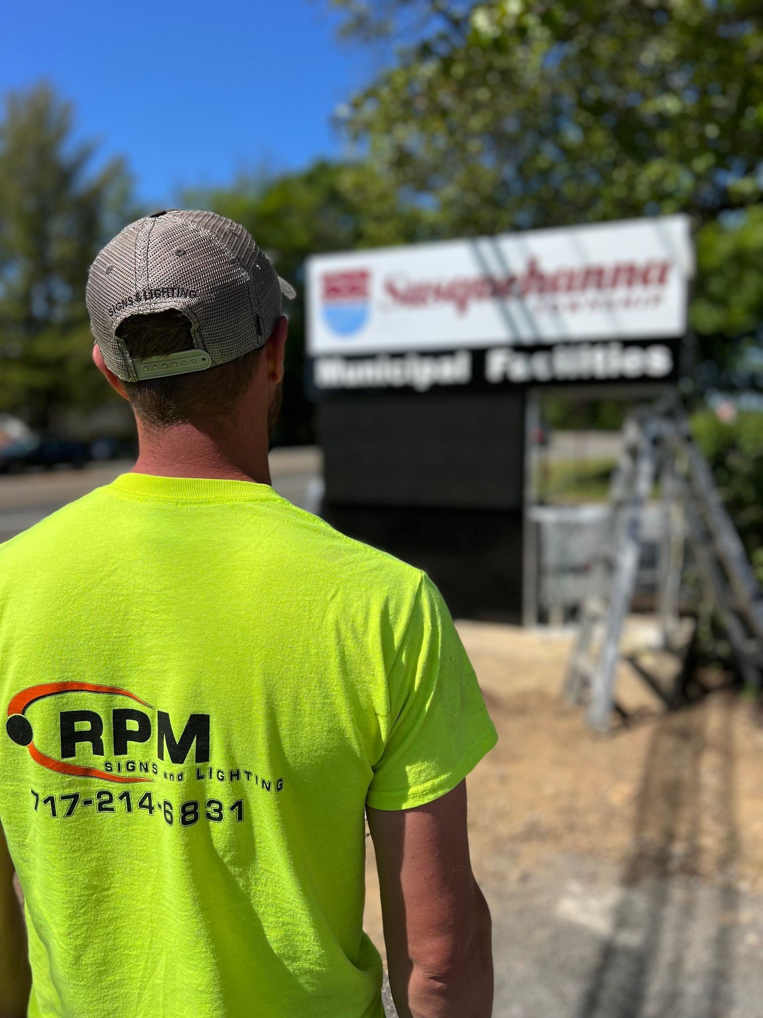 A man wearing a yellow shirt with the word rpm on the back is standing in front of a sign.