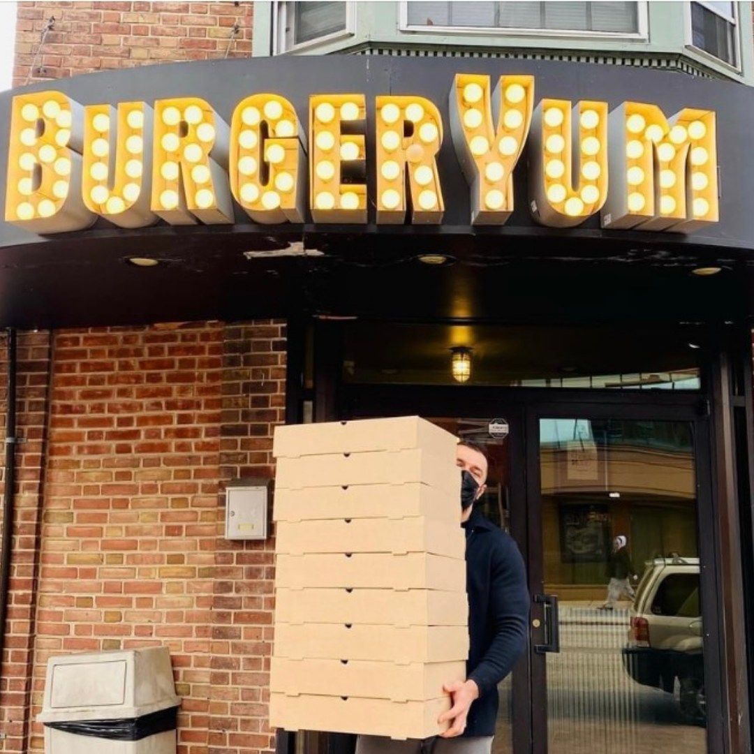 A man is carrying a stack of pizza in front of a burger yum sign