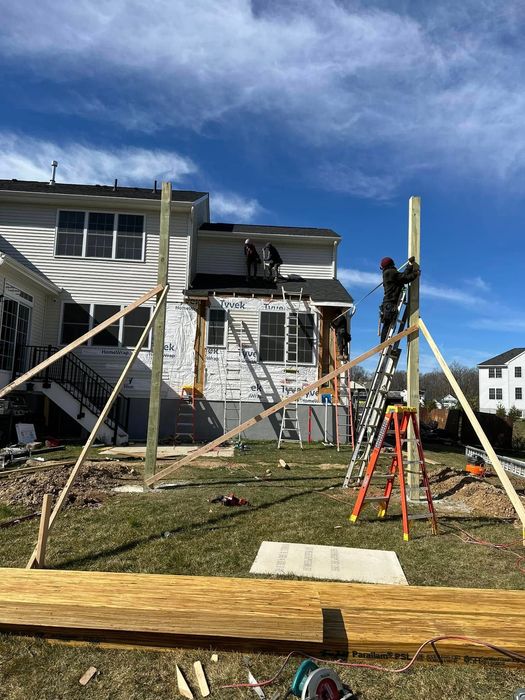 Construction workers building a structure, using ladders and bracing, next to a house under construction on a sunny day.