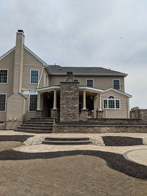 Back of a two-story beige house with stone patio and steps leading to yard on a cloudy day.