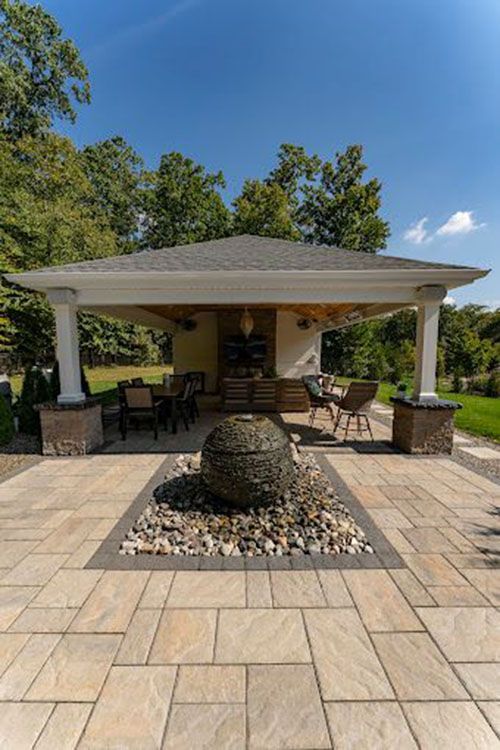 Outdoor patio with a pavilion, water feature, and dining area against a backdrop of trees and a blue sky.