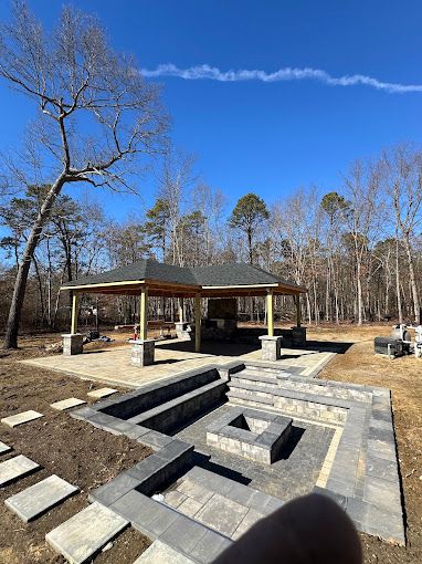 Outdoor patio with pavilion, fire pit, and seating area, under construction on a sunny day.