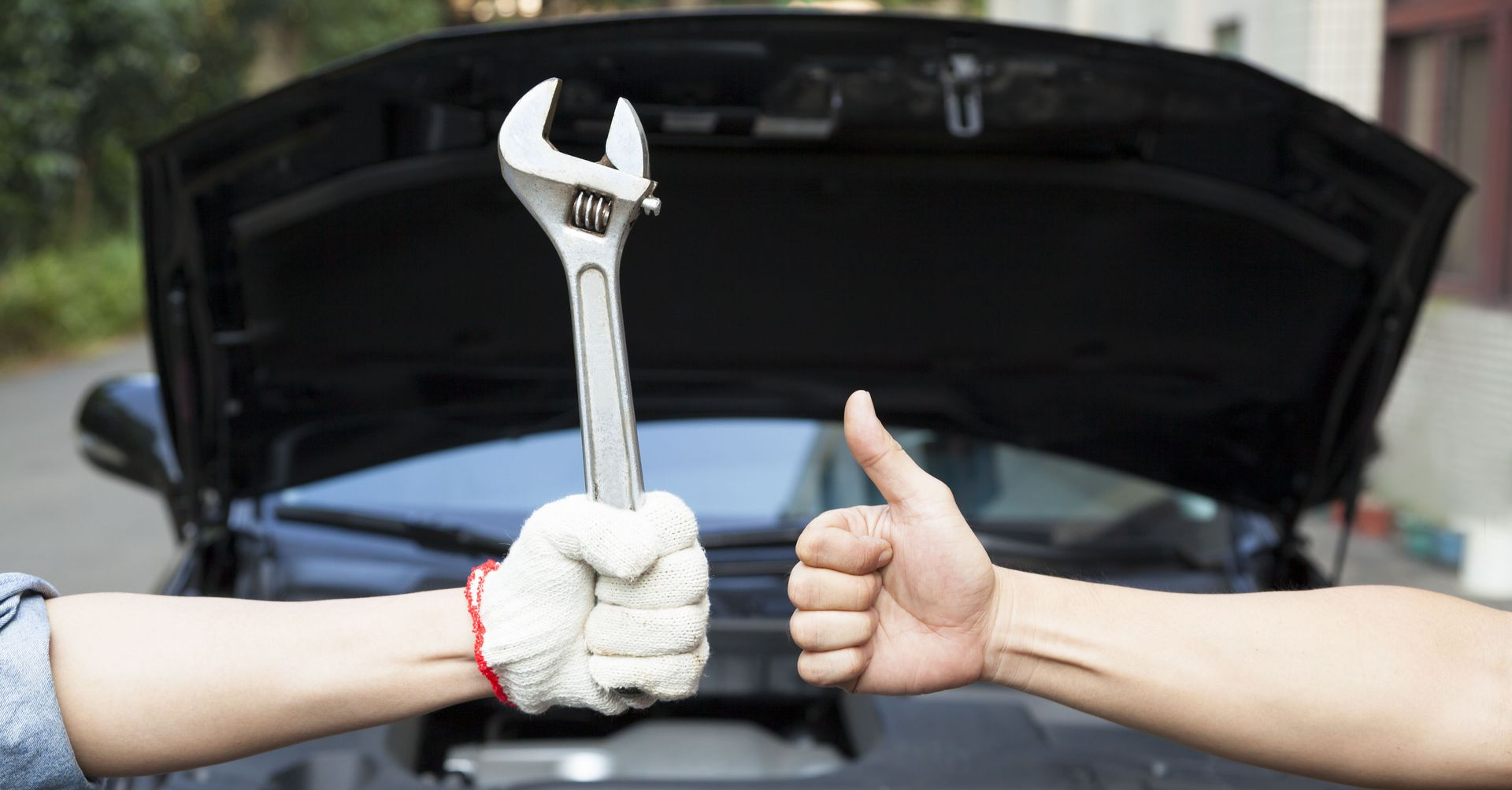 Hands holding wrench and giving thumbs up in front of an open car hood.
