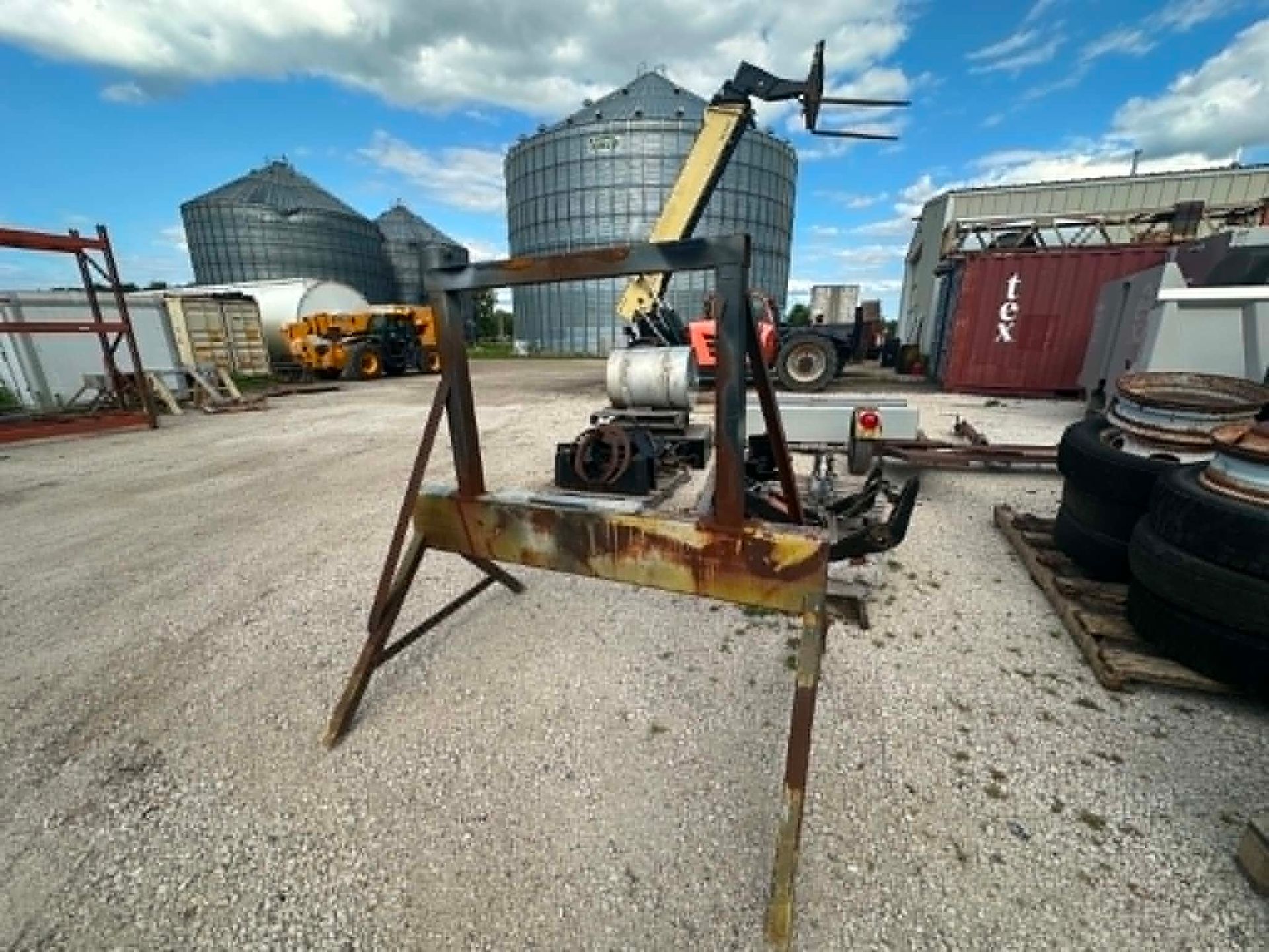 Rusty farm equipment in a gravel yard with grain silos and tractors in the background