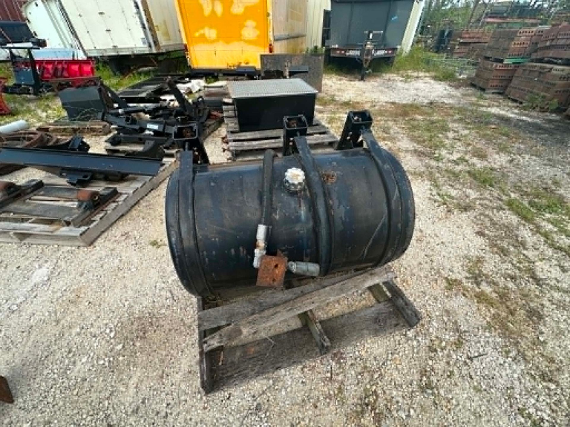 Black cylindrical tank on a wooden pallet in a gravel yard with scrap equipment nearby