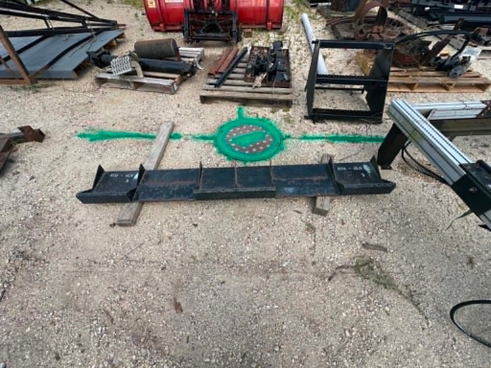 Outdoor metal fabrication area with steel parts, a green chain, and tools scattered on gravel.